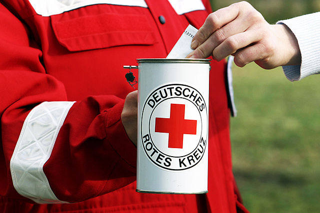 eine Person in roter Kleidung hält eine Spendendose in der Hand. Auf der Dose ist das DRK Logo abgebildet.