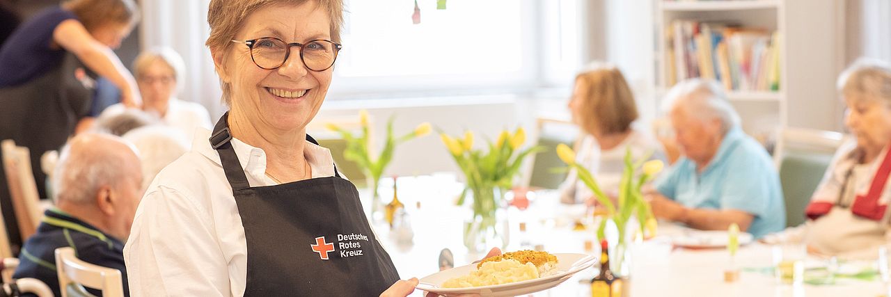 Senioren sitzen am gedeckten Tisch und eine Mitarbeiterin vom DRK serviert den Kuchen